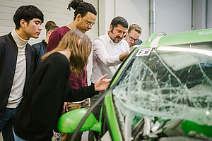 Professor inspiziert mit einer Gruppe von Studierenden ein gecrashtes Auto